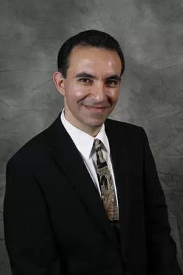 The image shows a man wearing a suit standing against a backdrop with a smile on his face.