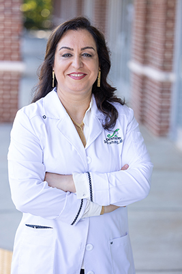 A woman in a white lab coat stands confidently against a brick wall with a sign behind her.