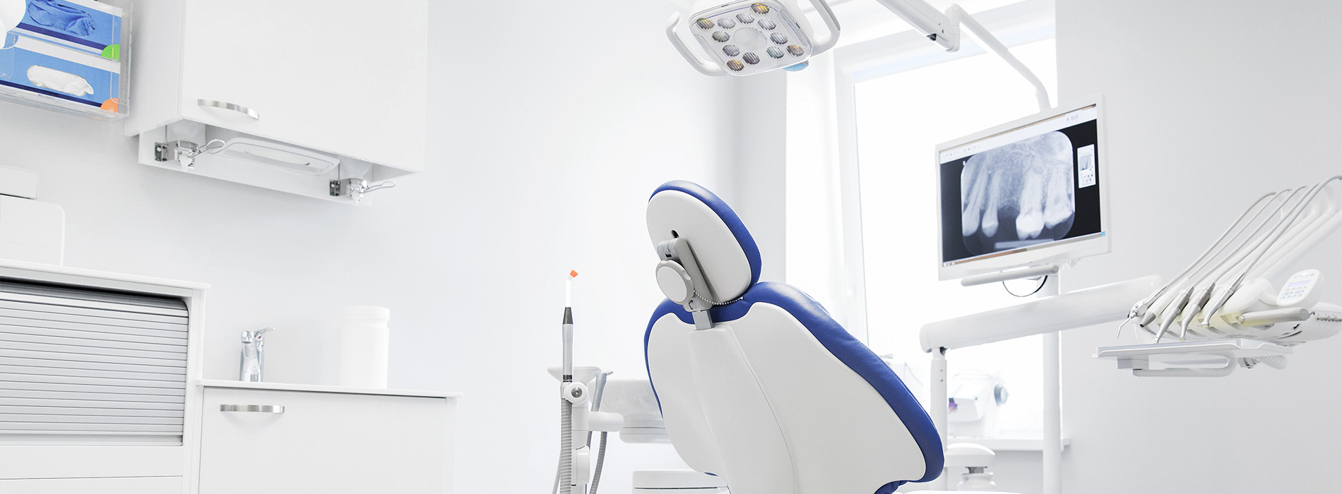 The image shows a modern dental office with advanced equipment, featuring a blue dental chair and various pieces of technology.