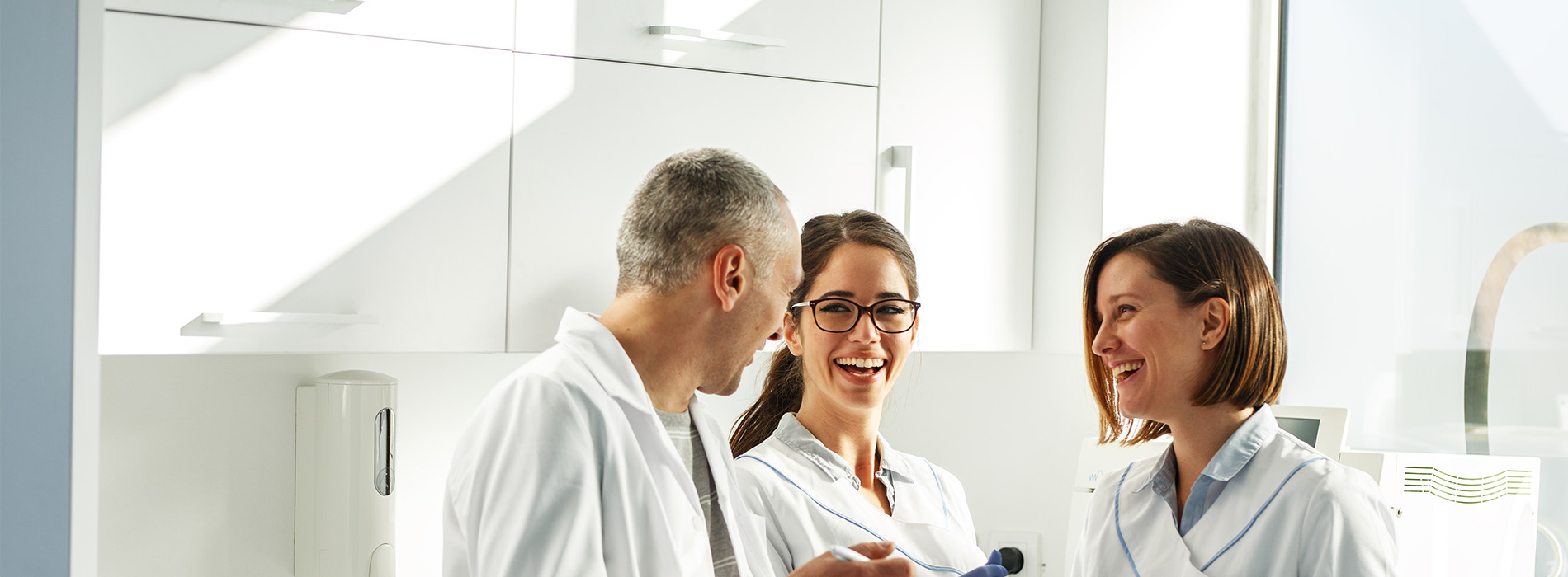 The image shows three people standing in front of a mirror, with two individuals on the left side of the frame and one on the right. They appear to be in an indoor setting, possibly a bathroom or a room with similar fixtures.