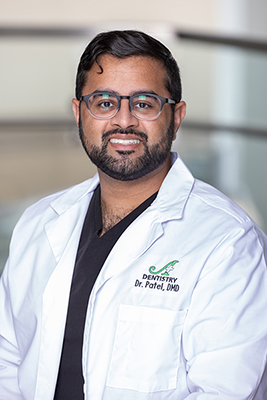 The image depicts a man wearing glasses, smiling at the camera, dressed in a white lab coat with a name tag, standing against a backdrop of a blurred indoor setting that appears to be an office environment.