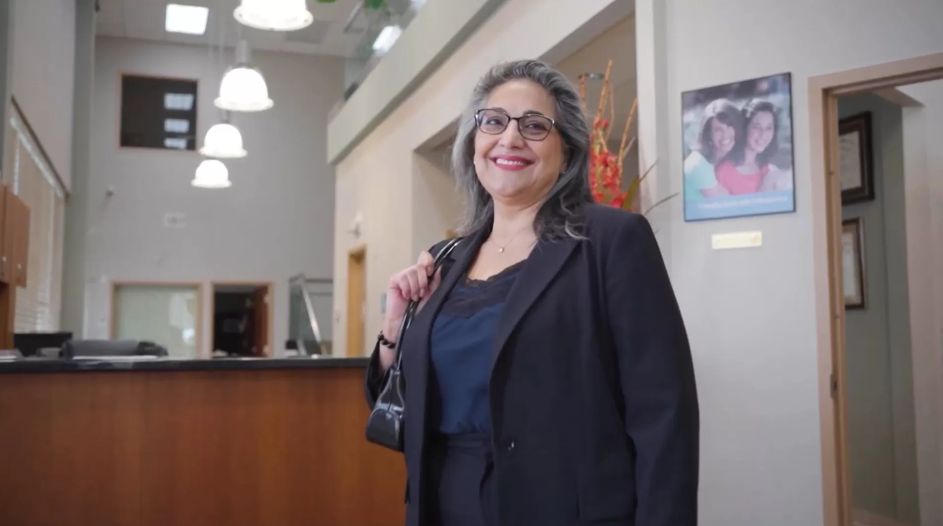 The image shows a woman standing inside a building, dressed in professional attire with a black jacket and glasses, posing for a photo with a smile. She has dark hair and appears to be middle-aged. In the background, there is a reception area with a desk, a sign, and a framed picture on the wall.