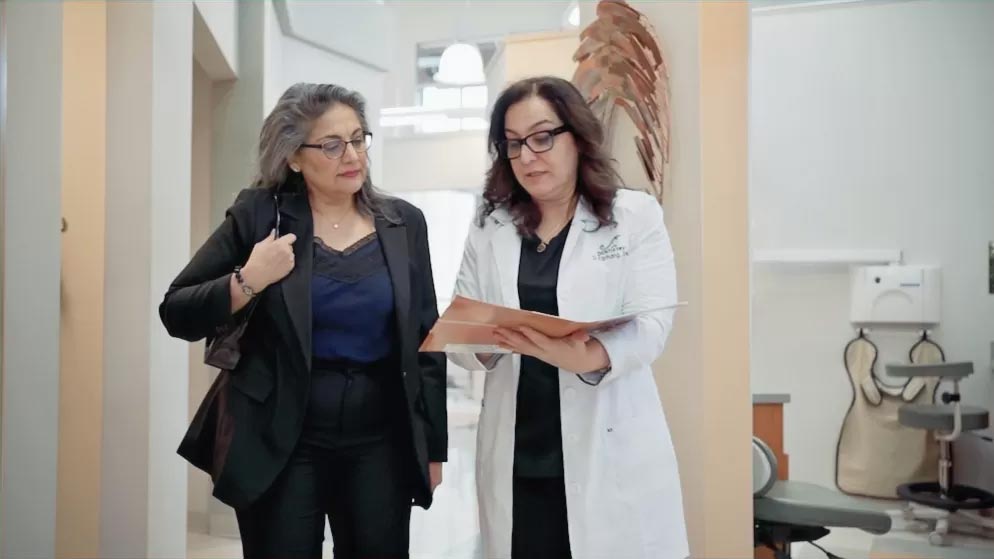 Two people standing in a room, with one person holding a clipboard, possibly discussing medical records or plans.