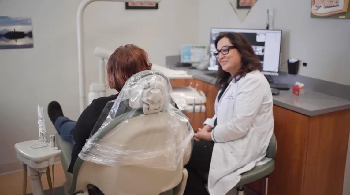 A dental hygienist is seated at a dental chair, adjusting protective plastic wrap over her face, while a patient sits in front of her, both in a dental office setting.
