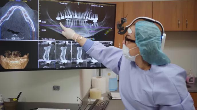 A dental professional in a surgical room pointing at a digital display with a 3D model of a mouth.