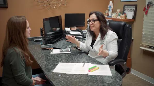 The image shows a woman sitting at a desk with a man standing in front of her, likely engaged in a conversation.