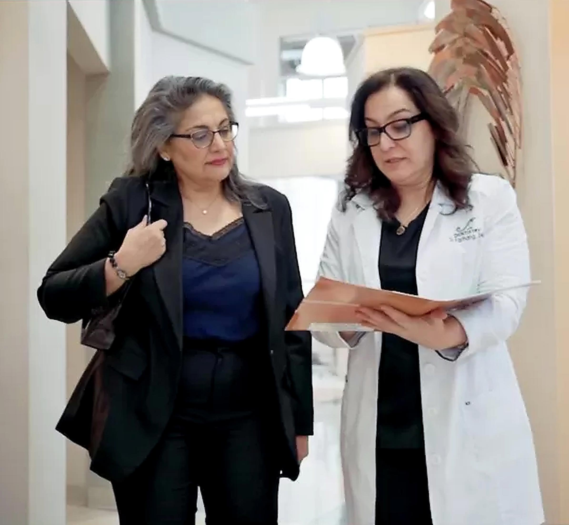 The image shows two individuals, likely professionals, standing together in an indoor setting with one person holding a document, possibly during a consultation or discussion.