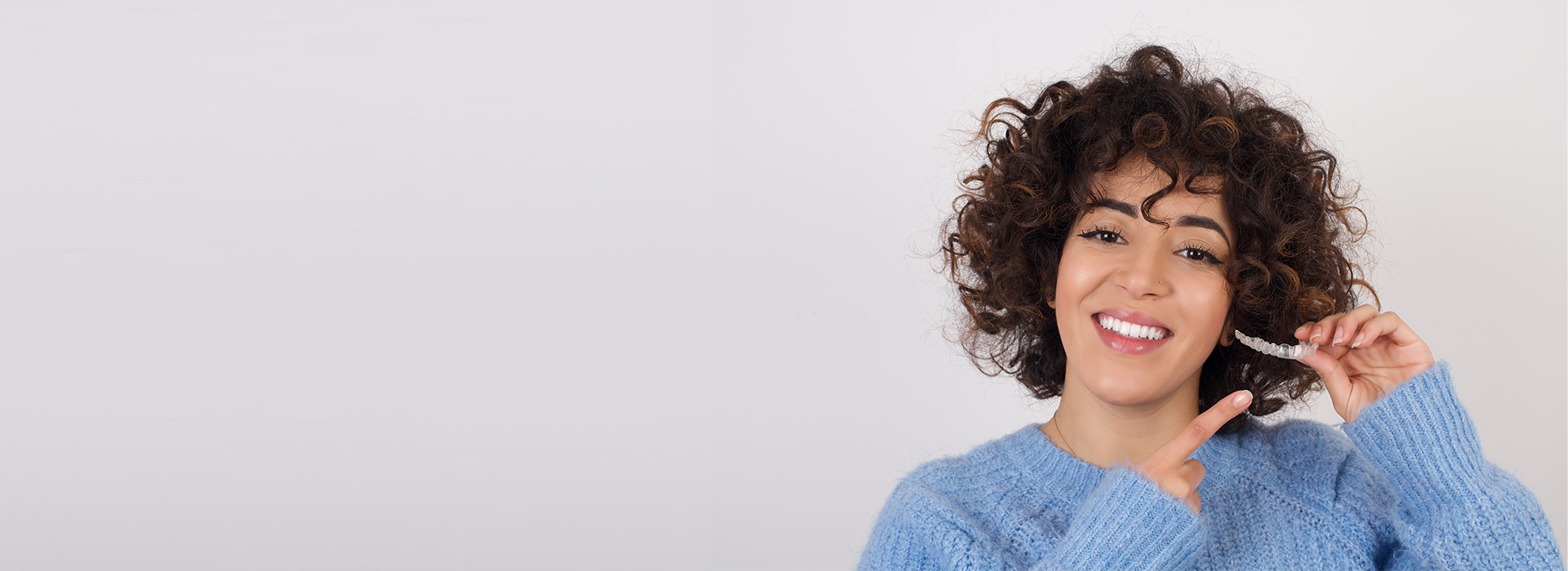 A person with curly hair brushing their teeth while smiling at the camera.