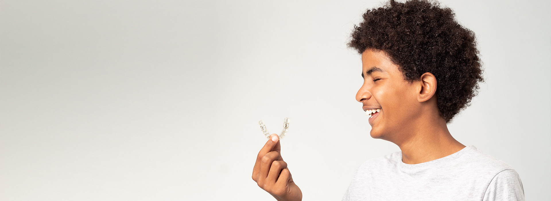 The image shows a person with curly hair holding a small object between their fingers, smiling at the camera, against a plain background.