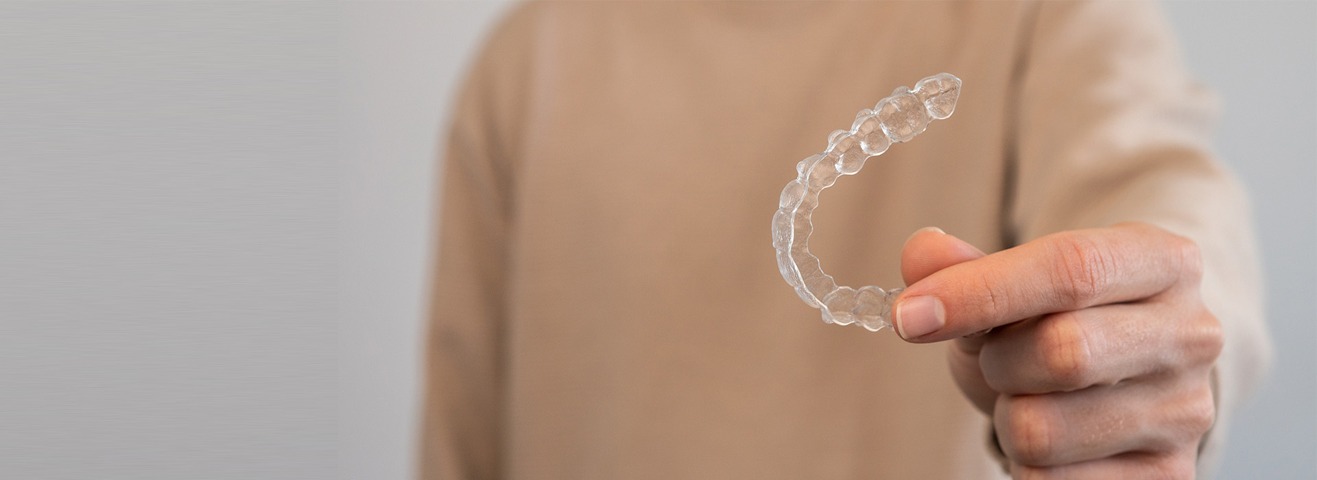 The image shows a person holding a clear plastic object with their left hand, which appears to be a ring or bracelet, against a blurred background that includes a portion of another person s upper body.