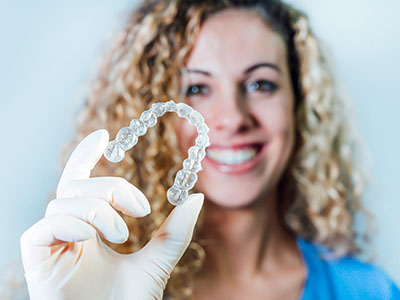 The image features a woman holding up a clear plastic container containing a set of teeth-shaped objects, presumably dental braces, against a white background.