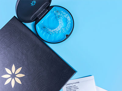 The image shows a close-up of a product packaging with a blue and white design, featuring two open boxes on a light blue surface, one containing a small item that appears to be a toothbrush, and another with a lid and a clear plastic container.