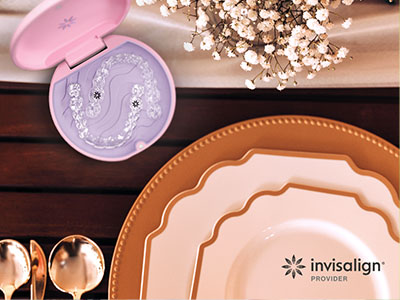 The image shows a table setting with various dishes and a pink makeup product box placed on top, set against a blurred background with a decorative item resembling a seashell.