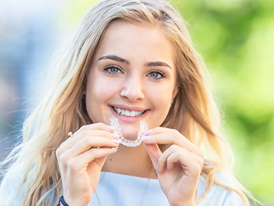The image features a young woman with light skin smiling at the camera while holding a toothbrush up to her teeth, which are visibly clean and white. She has long blonde hair and blue eyes, and she s wearing a casual top. There s a watermark on the bottom right corner of the image that reads  alt.