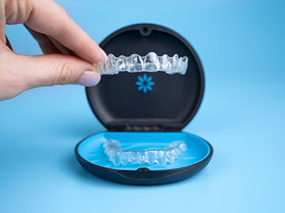 The image shows a person s hand holding an open box containing a clear plastic dental retainer with teeth-like ridges, against a blue background.