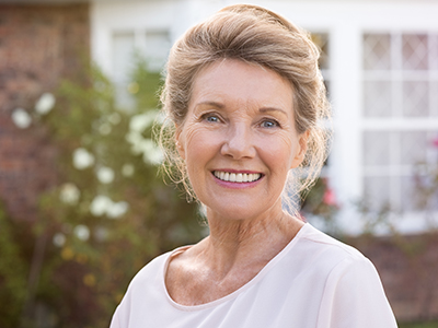 The image shows a woman standing outdoors in front of a house, smiling at the camera with a backdrop of greenery.