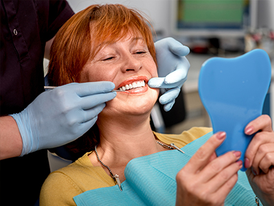 A woman sitting in a dental chair with her mouth open, receiving dental care, while holding up a blue toothbrush.