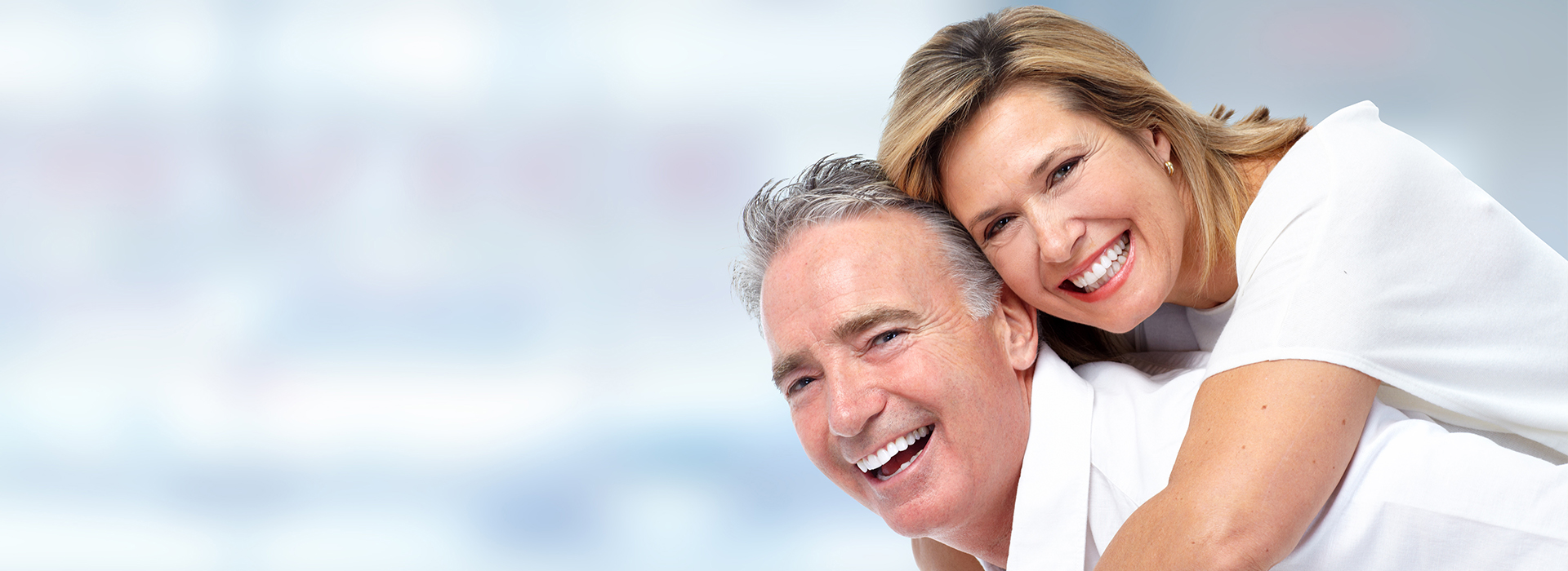 A photograph of a man and woman smiling and embracing, with the man holding the woman from behind, both wearing white tops and appearing happy.