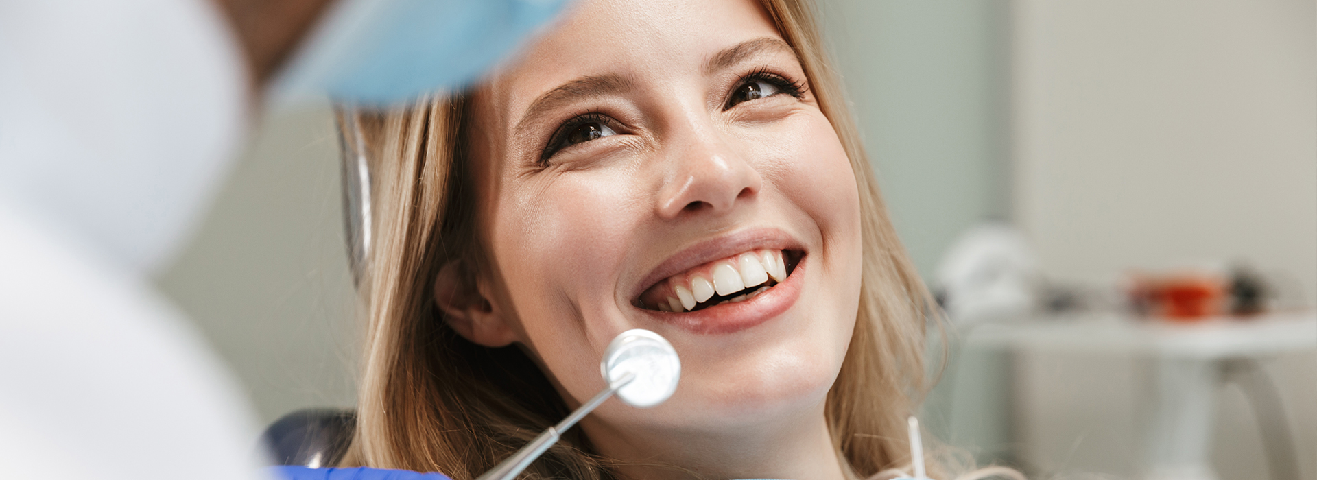 A young woman with blonde hair smiling at a dental mirror while receiving dental care from a professional.