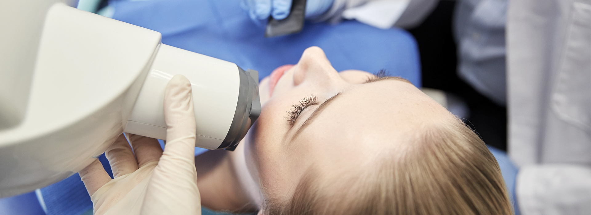The image shows a person receiving dental care with a handheld device, likely a dental mirror, being used by a professional wearing protective gloves and a face mask.