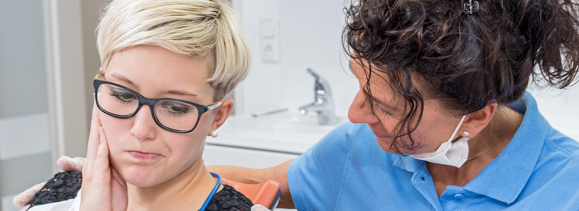 A woman with glasses sits in a dental chair while a dentist examines her teeth.