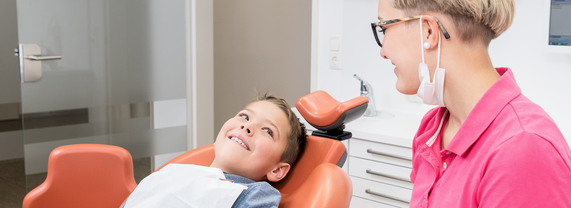 The image shows three people in an office setting  an adult male seated in a dental chair with his mouth open for a dental examination, a young child standing next to him wearing headphones, and a woman, presumably a dental professional, standing behind them both.