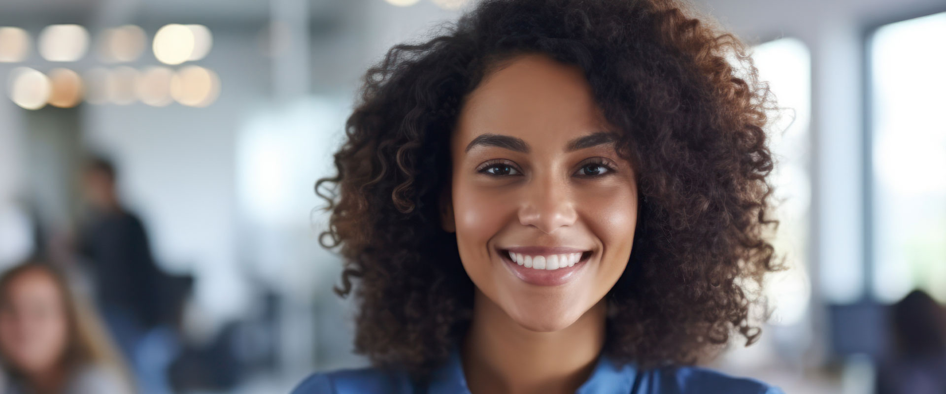 The image is a photograph of a smiling individual with curly hair, wearing a blue top, standing in an indoor setting with a blurred background that suggests a professional environment.