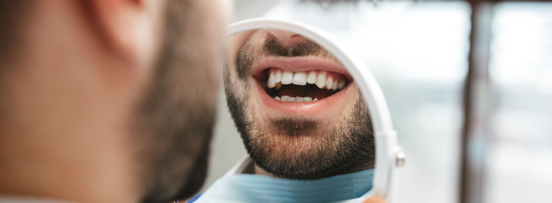 The image features a close-up of a man with an open mouth, showing his teeth, while he appears to be talking or laughing. He is wearing glasses and has facial hair. In the background, there s a partial view of another person who seems to be looking at him. The setting suggests an indoor environment, possibly a dental office or a similar professional space.