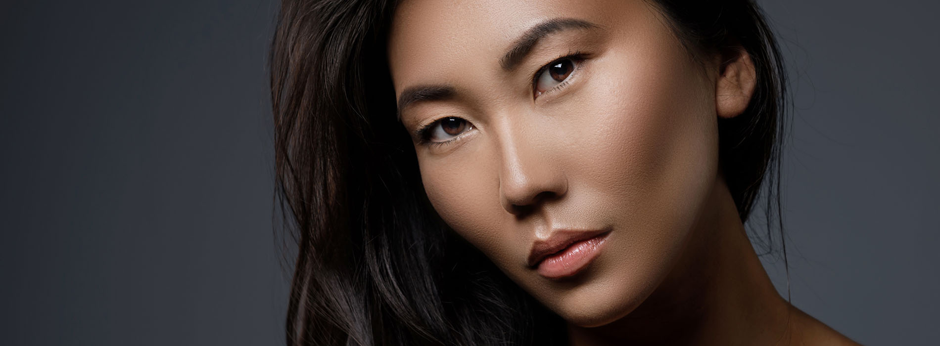 The image features a woman with long dark hair and fair skin, wearing makeup and looking directly at the camera, set against a dark background.
