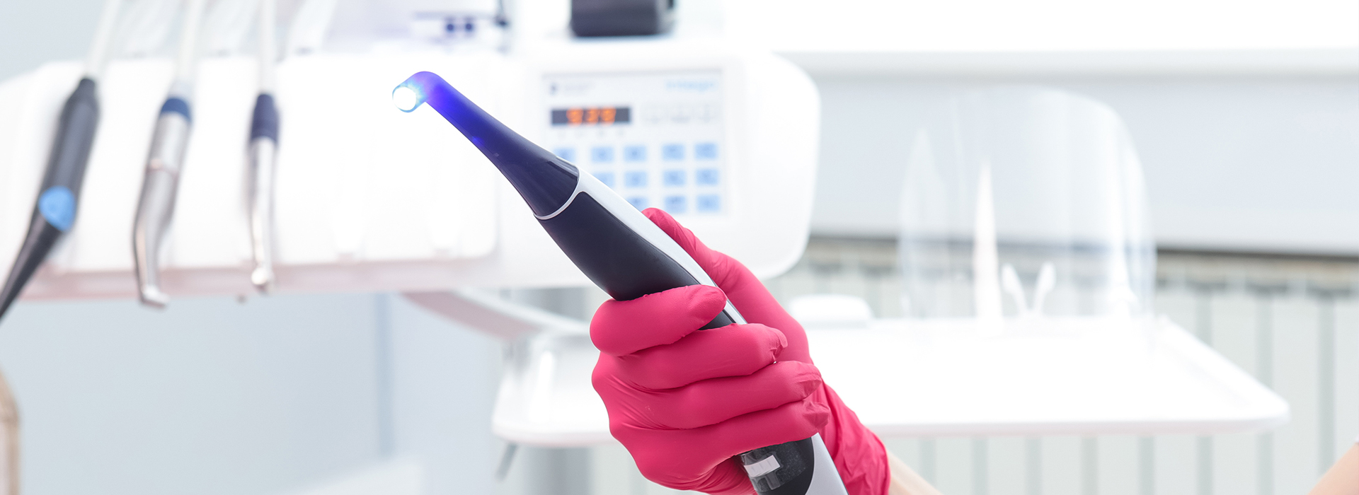 The image shows a close-up view of a person s hand holding an electronic device with a blue light, wearing pink gloves, and standing in a laboratory setting with various equipment in the background.