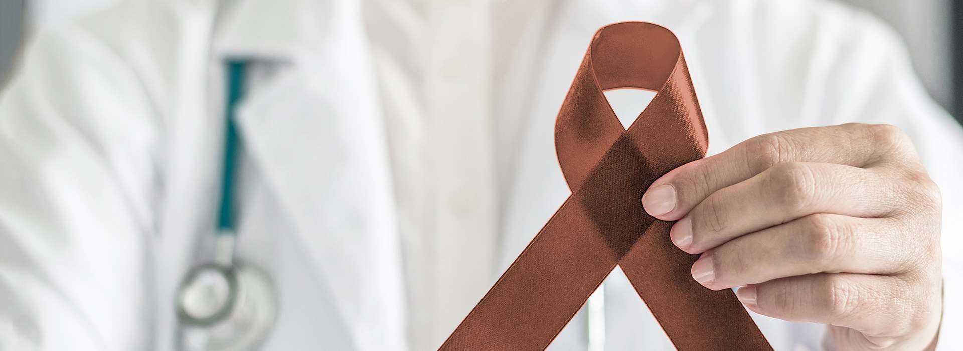 The image shows a hand holding a red ribbon with a white cross symbolizing medical aid, placed over an image of a person wearing a stethoscope, suggesting a theme related to healthcare and possibly breast cancer awareness.
