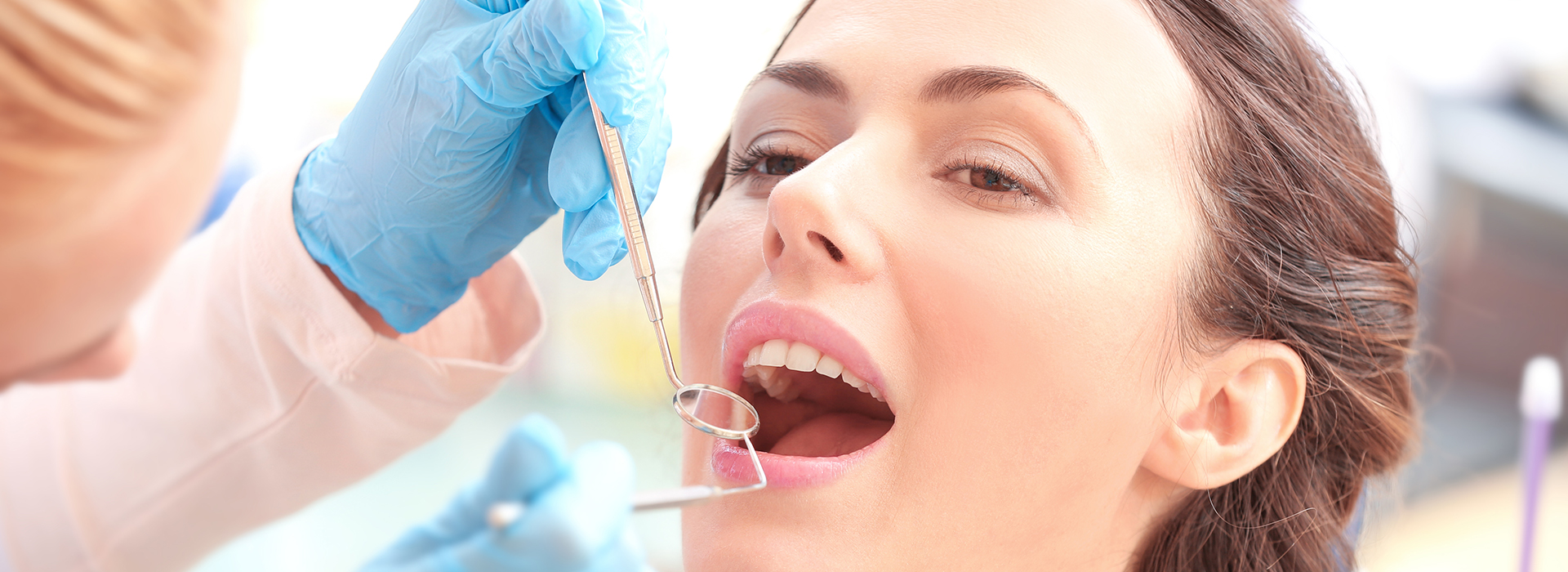 The image shows a woman with her mouth open, receiving dental care from a professional in a clinical setting, with visible dental tools such as a drill and gauze being used on her teeth.