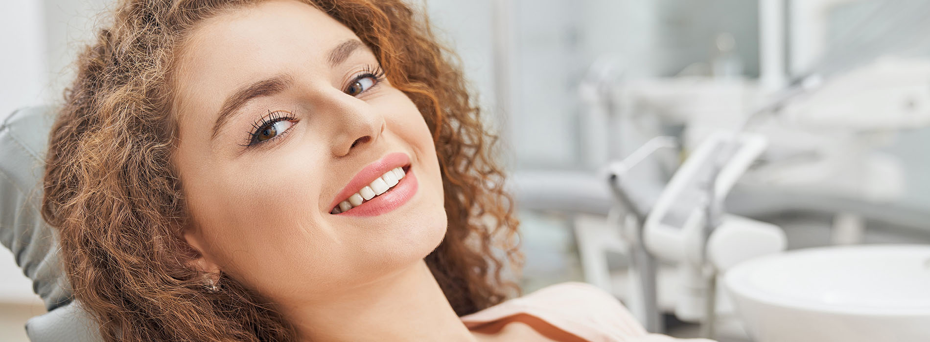 The image shows a smiling woman with curly hair sitting in a dental chair, looking directly at the camera, with the background featuring dental equipment and a professional setting.