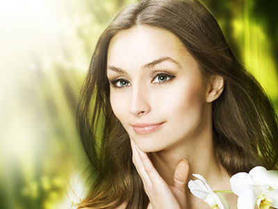 A woman with long hair looking directly at the camera, showcasing her beauty with a soft smile against a blurred background featuring green foliage.