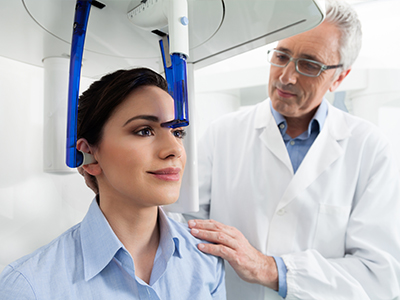 The image shows a woman seated in front of a large piece of medical equipment with a man standing behind her, both inside a clinical setting.