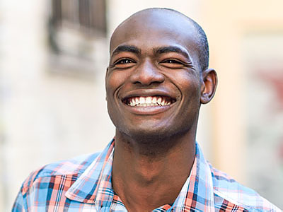 The image shows a smiling man with short hair, wearing a plaid shirt, standing outdoors during the daytime.