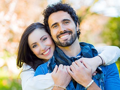 A man and woman are embracing each other outdoors  the man has facial hair, both individuals appear happy, and they are dressed casually.