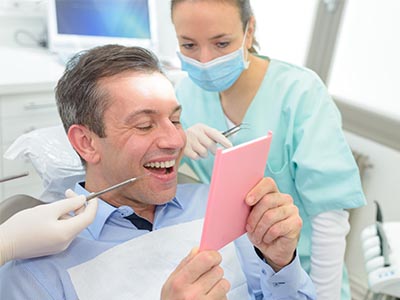 The image shows a man sitting in a dentist s chair with a tablet displaying a photo of himself, smiling broadly, while a dental professional looks on, possibly during a dental appointment or procedure.