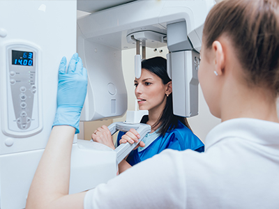A woman wearing a blue jacket stands behind a large medical imaging machine, while another person is seated in front of it, possibly for an examination.