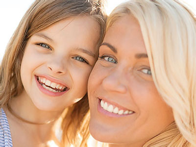 The image shows two individuals, an adult woman and a young child, smiling at the camera against a light background with clear skies.