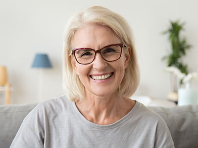 The image features a woman with blonde hair smiling at the camera, wearing glasses and a light blue top. She appears to be indoors, possibly in a living room setting, with a neutral background that includes a lamp and part of a couch.