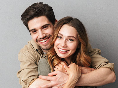 This is a photograph of a man and a woman embracing each other with smiles on their faces, both looking directly at the camera.