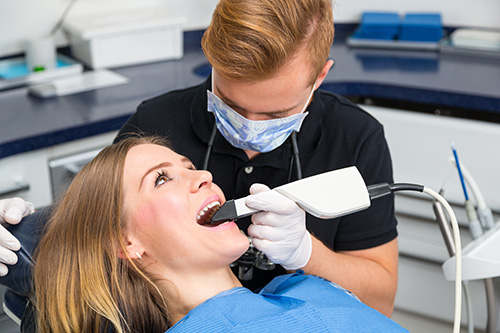 The image shows a dental hygienist performing a cleaning procedure on a patient s teeth while standing behind her, with both individuals wearing personal protective equipment.