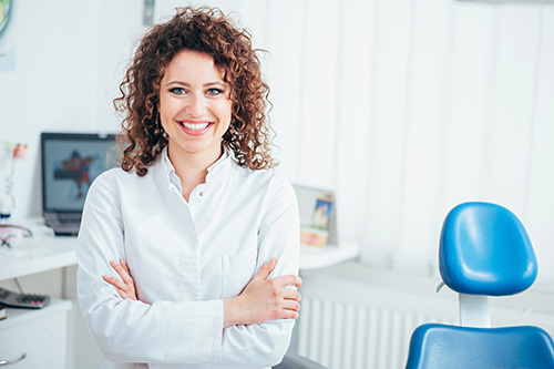 A woman with curly hair is standing in an office setting, smiling at the camera while leaning against her desk.