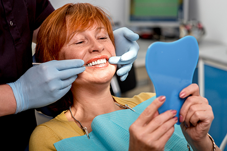 A woman with red hair is sitting in a dental chair, holding up a blue toothbrush-shaped object, smiling broadly while looking at the camera.