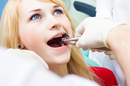 The image depicts a woman receiving dental care, with a dental professional performing a procedure on her teeth while she holds her mouth open and appears to be seated in a dental chair.