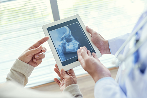 The image shows two hands holding a tablet displaying an X-ray, with a person in a white coat pointing at the screen, suggesting medical consultation or diagnosis being discussed.