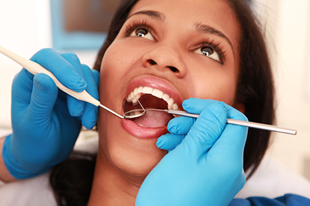A woman undergoing dental treatment, with her mouth open wide, receiving care from a dentist wearing blue gloves and using dental tools.