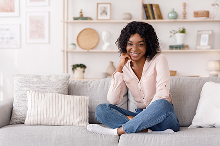 A woman sitting on a couch with her legs crossed, smiling at the camera, wearing a pink blouse and blue jeans, in a room decorated with white walls, framed pictures, and plants.