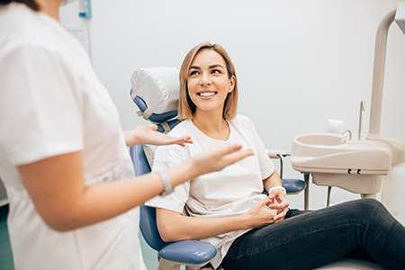 A dental hygienist is seated at a dental chair, smiling at a woman who is seated in front of her, both wearing white shirts, with another person standing behind them.